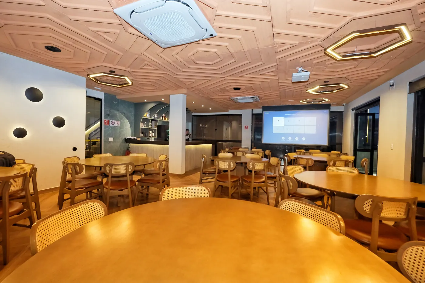 Sala do 2º andar montada com mesas redondas, telão ligado ao fundo e bar ao lado.